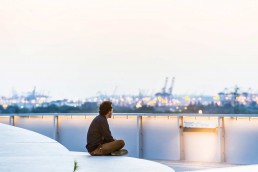ein junger Mann sitzt im Schneidersitz auf einer skulpturalen Fläche und guckt auf die Kräne und Lichter im Hafen