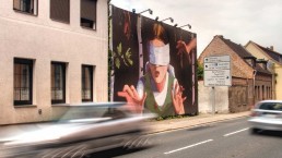 Autos fahren auf einer Straße, in der eine Brachfläche mit einer Plakatwand bespielt wird, darauf ein gemaltes Mädchen mit verbundenen Augen