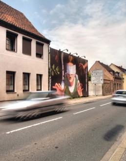 Autos fahren auf einer Straße, in der eine Brachfläche mit einer Plakatwand bespielt wird, darauf ein gemaltes Mädchen mit verbundenen Augen