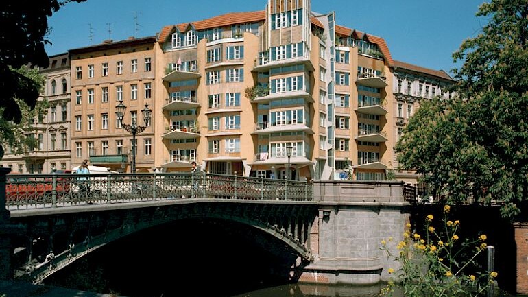 im Vordergrund ein Kanal mit einer historischen Brücke, im Hintergrund ein großes Eckgebäude einer Blockrandbebauung mit geschwungenen Balkonen und Erkern