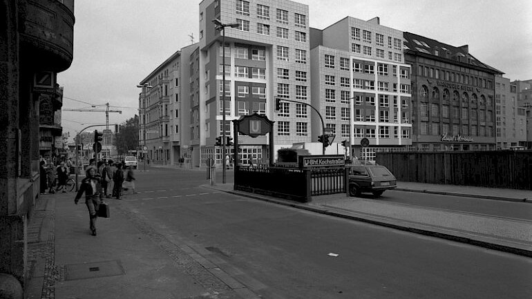 im Vordergrund eine Straße und ein U-Bahn-Aufgang, im Hintergrund ein Eckgebäude mit gerasterter Fassade und ohne Dachaufbau