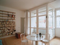 ein helles Zimmer mit Bücherregal und Bistrotisch und Stühlen, im Hintergrund eine noch nicht möblierte Loggia