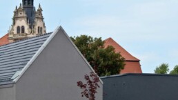 zwei neue graue Gebäude, eines mit Satteldach, eines mit Flachdach, im Hintergrund der Turm einer historischen Kirche