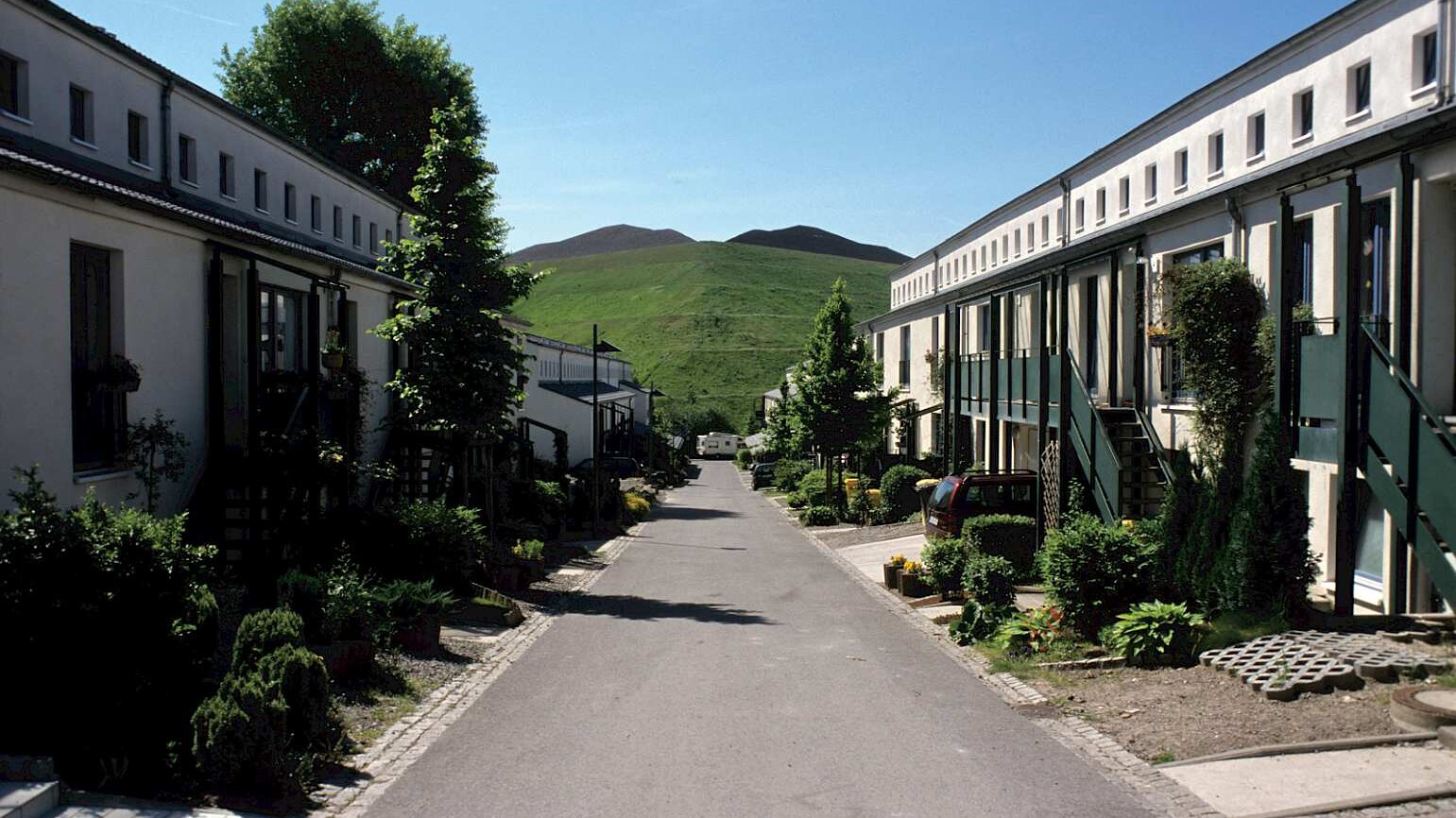 eine kleine Wohnstraße führt auf einen grünen Hügel, links und rechts helle Reihenhäuser mit Vorgärten