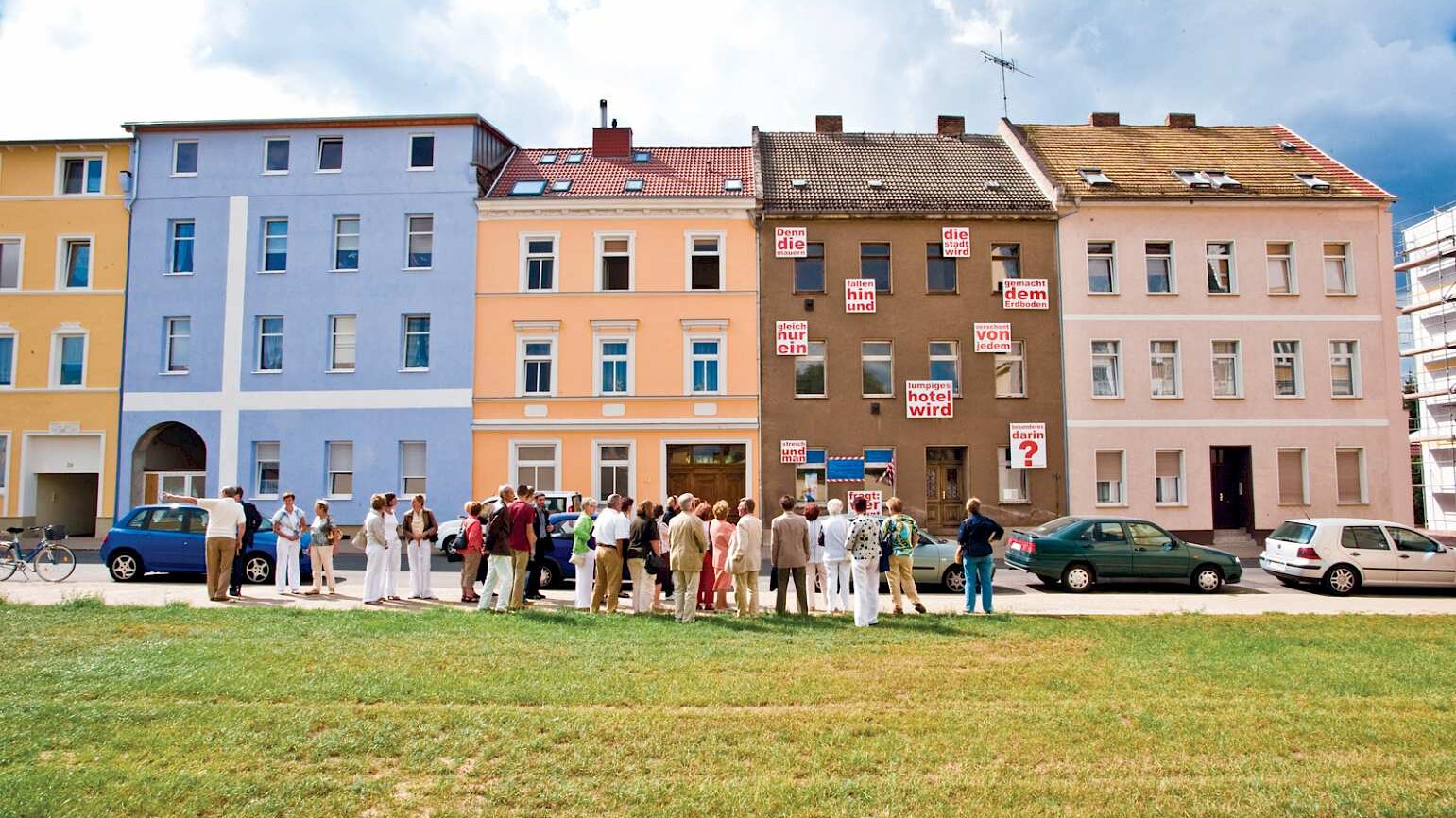 eine Gruppe von Menschen steht vor einer Reihe farbiger, dreistöckiger Altstadthäuser, im Vordergrund eine rasenbewachsene Brachfläche