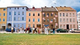 eine Gruppe von Menschen steht vor einer Reihe farbiger, dreistöckiger Altstadthäuser, im Vordergrund eine rasenbewachsene Brachfläche