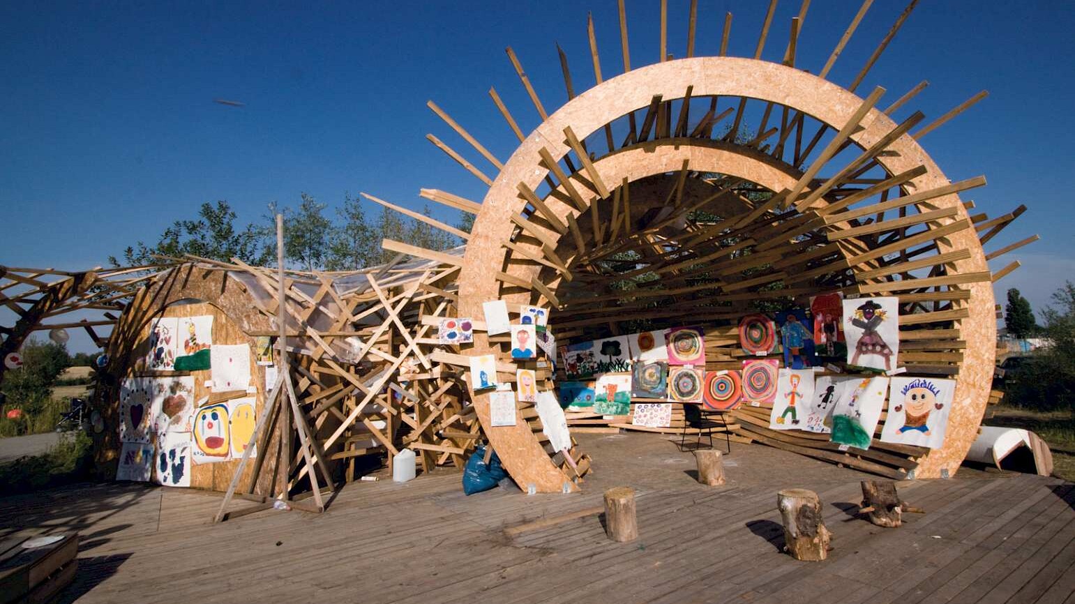 in einer runden, offenen Holzkonstruktion hängen großformatige, von Kindern gemalte Bilder