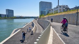 Spazierende und Radfahrende nutzen den Uferweg am Wasser, im Hintergrund modernde Bürohäuser
