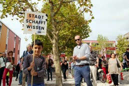 ein Junge hält ein Schild mit der Aufschrift „Stadt schafft Wissen“ hoch, im Hintergrund weitere Menschen