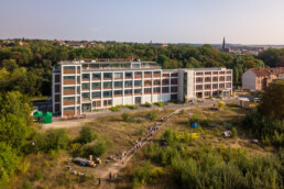 ein fein strukturiertes Fabrikgebäude in grüner Landschaft mit einer Gruppe von Menschen im Vordergrund